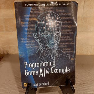 May include: A book titled "Programming Game AI by Example" by Mat Buckland. The cover features a wireframe human head and technical terms like "Path," "Logic," and "Memory." The book is displayed on a black metal stand.