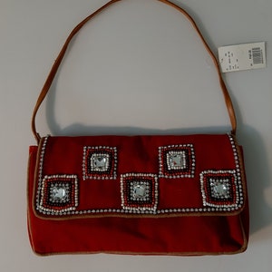 May include: A red handbag with a brown leather strap. The flap is decorated with square patterns of white, red, and clear beads. The bag has a brown trim and a tag attached.