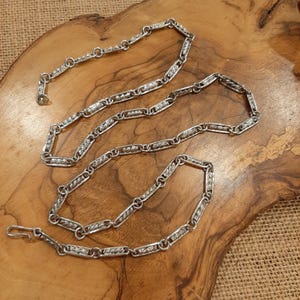 May include: A silver-toned metal chain necklace featuring a series of elongated rectangular links with a textured, twisted pattern, connected by small circular jump rings. The chain includes a simple hook-style clasp at one end for closure.