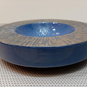 May include: A blue ceramic dish with a textured rim and a smooth, slightly lighter blue interior. The dish is round and has a shallow depth.