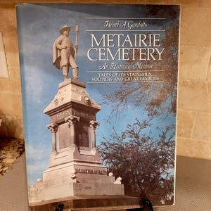 May include: A book titled "Metairie Cemetery: An Historical Memoir" by Henri A. Gandolfo. The cover features a statue atop a monument against a blue sky. The book's subtitle reads "Tales of Its Statesmen, Soldiers and Great Families."