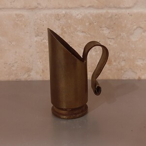 May include: A small brass vessel shaped like a coal scuttle, featuring a cylindrical body with a slanted top opening and a curved handle attached to the side. The metal has a muted, aged finish with visible signs of wear and oxidation throughout.