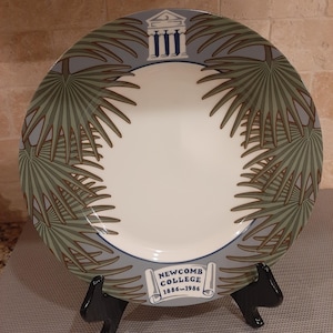 May include: A decorative plate featuring a white center and a gray border adorned with green palm fronds. The plate includes a blue and white architectural design and the text "NEWCOMB COLLEGE 1886-1986" on a banner. The plate is displayed on a black stand.