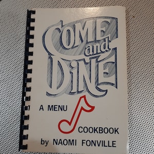 May include: A spiral-bound cookbook titled "Come and Dine" with blue and red accents. The cover features the title in a stylized font, a red musical note, and the author's name, Naomi Fonville. The cookbook is a menu cookbook.