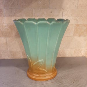 May include: A flared ceramic vase with a scalloped rim and vertical fluted panels. The upper portion is a matte teal green, transitioning into a warm brown base featuring a raised relief design of three stylized leaves wrapping around the bottom.