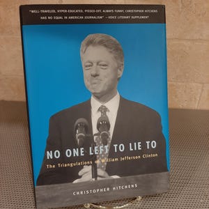 May include: A hardcover book titled "NO ONE LEFT TO LIE TO" by Christopher Hitchens. The book cover features a portrait of William Jefferson Clinton against a blue background. The book is displayed on a silver stand.