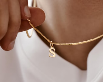 May include: Gold-colored necklace with a small pendant in the shape of the letter "S". The chain has a textured appearance. The necklace is being held by a person wearing a white shirt.