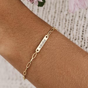Könnte beinhalten: Ein goldfarbenes Kettenarmband mit einem kleinen, rechteckigen Metallanhänger, der mit dem Wort "sababa" in einer einfachen Kleinbuchstabenschrift graviert ist. Das Armband besteht aus länglichen ovalen Gliedern und einem passenden Verschluss.