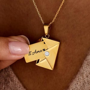 Puede incluir: Un collar dorado con un colgante en forma de sobre. El colgante tiene un pequeño acento blanco redondo y las palabras "Ti Amo" son visibles en la tarjeta extraíble. El collar se lleva alrededor del cuello.