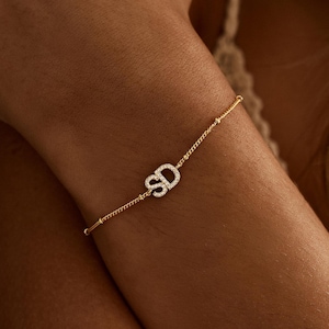 May include: A delicate gold bracelet with the initials "SD" encrusted with small, sparkling stones. The bracelet is strung with tiny gold beads and is worn on a wrist.