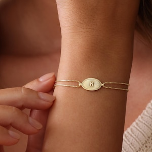 May include: A gold-toned bracelet featuring a delicate double chain and a central oval charm. The charm has a textured border and displays the capital letter N in the centre. The bracelet is worn on a wrist, showcasing its fine metal construction.