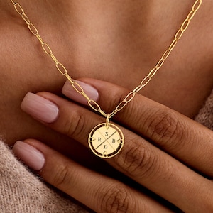 Peut inclure: Un collier doré avec un pendentif circulaire portant les lettres S, R, B et D. Le pendentif est suspendu à une chaîne délicate. Le collier est présenté sur la poitrine d'une personne.