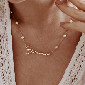 Puede incluir: Un collar de tono dorado con una cadena fina, pequeñas perlas blancas espaciadas uniformemente y un colgante central con el nombre Eleana en letra cursiva. El collar se apoya sobre una prenda de vestir de tela blanca texturizada.