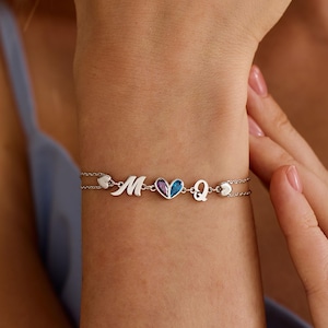 May include: A silver-toned double chain bracelet featuring a central heart charm split into purple and blue sections, flanked by the letters M and Q in a cursive script, and finished with small heart accents on each side of the chain links.