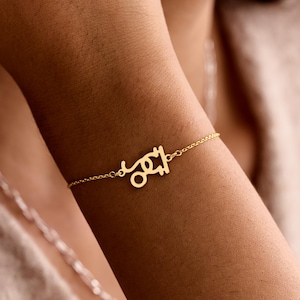 May include: A delicate gold-colored bracelet featuring a Capricorn zodiac symbol. The bracelet is attached to a thin chain and is worn on a wrist. The background is blurred, with a focus on the jewelry.