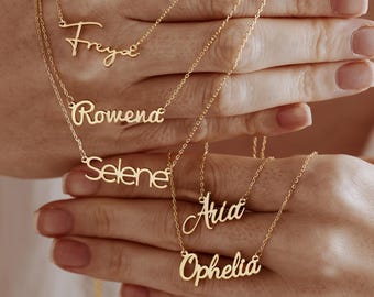 May include: Gold-colored name necklaces with delicate chains. The names "Freya", "Rowena", "Selene", "Aria", and "Ophelia" are displayed in cursive script. The necklaces are held up against a white background.