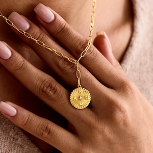 May include: A gold-colored necklace with a circular pendant featuring a starburst design and engraved letters. The necklace has a delicate chain and is being worn. The pendant hangs from a shorter chain.