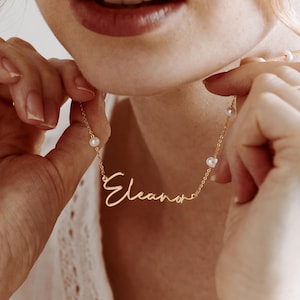May include: A gold-toned metal necklace featuring a delicate chain with small, evenly spaced white pearls and a central script-style nameplate pendant that reads "Eleanor" in a cursive font, suspended from a thin, polished metallic link chain.