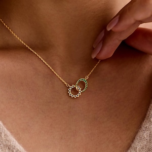 May include: A gold-colored necklace featuring two interlocking circles. One circle is adorned with blue gemstones, the other with green gemstones. The necklace is suspended from a delicate gold chain.