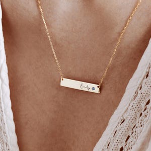 Könnte beinhalten: Eine goldfarbene Halskette mit einem rechteckigen Anhänger. Der Anhänger trägt den Namen "Emily" in Schreibschrift und einen kleinen blauen Edelstein. Die Halskette wird vor einem weißen Hintergrund gezeigt.