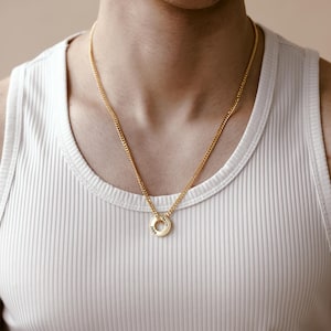 Op de afbeelding: Een goudkleurige ketting met een cirkelvormige hanger met gegraveerde tekst. De hanger hangt aan een gourmette ketting. De persoon draagt een witte geribbelde tanktop.