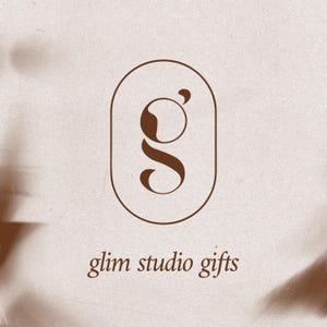 May include: A logo for "glim studio gifts" features a stylized brown letter "g" inside a brown oval frame. The text "glim studio gifts" is written in a brown, elegant font below the logo, set against a beige background with soft shadows.
