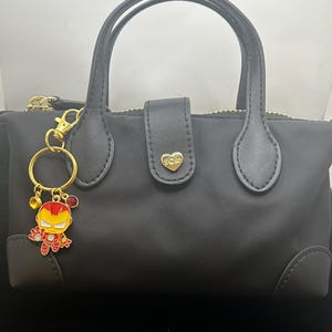 K&ouml;nnte beinhalten: Schwarze Handtasche mit goldenem Rei&szlig;verschluss und Griffen. Ein goldener Schl&uuml;sselanh&auml;nger mit einer Iron Man-Cartoonfigur ist angebracht. Der Schl&uuml;sselanh&auml;nger hat rote und gelbe Akzente. Die Tasche hat einen herzf&ouml;rmigen goldenen Verschluss.