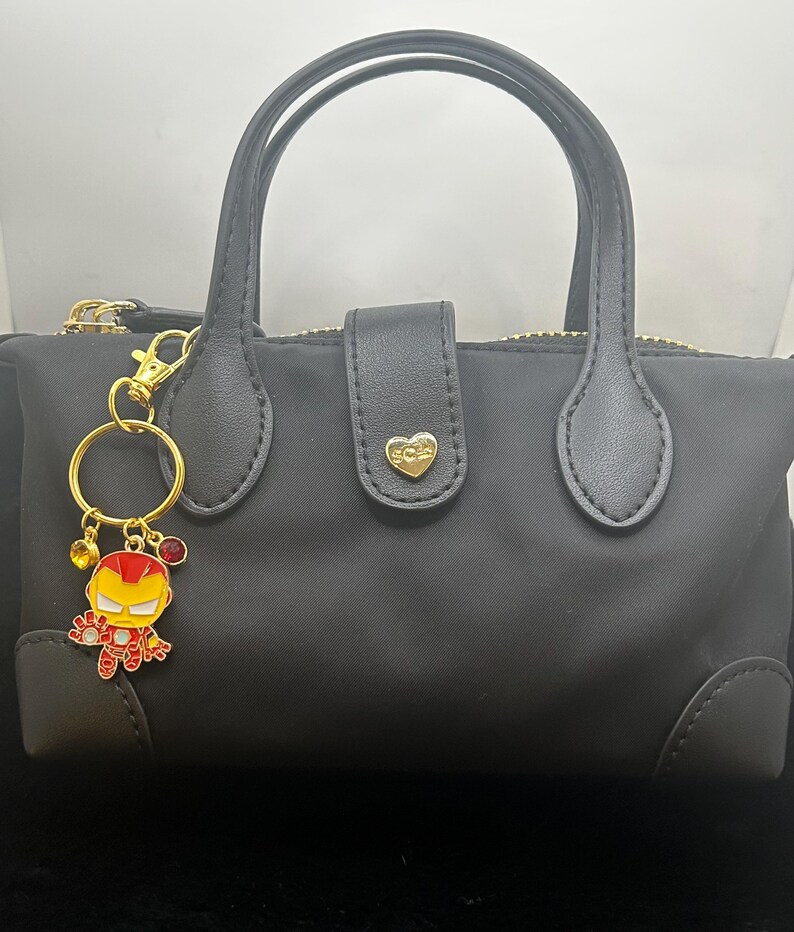 K&ouml;nnte beinhalten: Schwarze Handtasche mit goldenem Rei&szlig;verschluss und Griffen. Ein goldener Schl&uuml;sselanh&auml;nger mit einer Iron Man-Cartoonfigur ist angebracht. Der Schl&uuml;sselanh&auml;nger hat rote und gelbe Akzente. Die Tasche hat einen herzf&ouml;rmigen goldenen Verschluss.