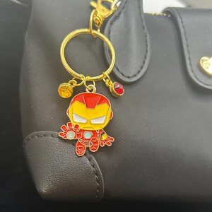 K&ouml;nnte beinhalten: Ein goldfarbener Schl&uuml;sselanh&auml;nger mit einem Iron Man-Cartoon-Anh&auml;nger. Der Anh&auml;nger ist rot, gelb und wei&szlig; mit einem goldfarbenen Ring und Verschluss. Der Schl&uuml;sselanh&auml;nger hat drei kleine dekorative Anh&auml;nger: einen gelben Edelstein, einen roten Edelstein und einen goldfarbenen Anh&auml;nger.