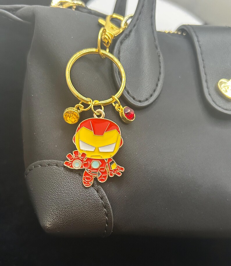 K&ouml;nnte beinhalten: Ein goldfarbener Schl&uuml;sselanh&auml;nger mit einem Iron Man-Cartoon-Anh&auml;nger. Der Anh&auml;nger ist rot, gelb und wei&szlig; mit einem goldfarbenen Ring und Verschluss. Der Schl&uuml;sselanh&auml;nger hat drei kleine dekorative Anh&auml;nger: einen gelben Edelstein, einen roten Edelstein und einen goldfarbenen Anh&auml;nger.