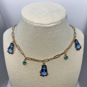 Puede incluir: Un collar de cadena dorada con múltiples colgantes. Los colgantes incluyen figuras de dibujos animados azules y pequeñas piedras preciosas azules redondas. El collar se exhibe en un busto de maniquí blanco.