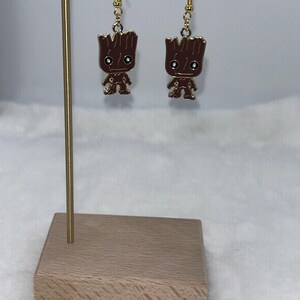 May include: A pair of gold-colored dangle earrings featuring brown Groot figures. The earrings are displayed on a gold-colored stand with a wooden base. The Groot figures have a brown body and gold accents.