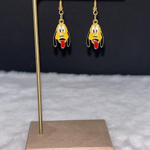 May include: A pair of gold-colored dangle earrings featuring a yellow cartoon dog with black accents and a red tongue. The earrings are displayed on a gold-colored stand with a wooden base, against a black background.