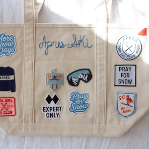 Può includere: Borsa tote color crema con diverse toppe ricamate. Le toppe includono frasi come "More Snow Days", "Apres Ski" e "Pray for Snow". Altre toppe presentano grafiche e testo a tema sci. La borsa ha due manici.