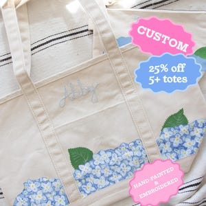 Może przedstawiać: Kremowa torba typu tote z niebieskim kwiatowym wzorem hortensji. Torba ma przednią kieszeń i uchwyty. Słowo "July" jest wyhaftowane z przodu. Ręcznie malowane i haftowane.