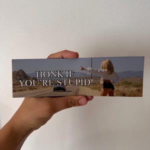 May include: Rectangular sticker with a desert road scene and the text "HONK IF YOU'RE STUPID!". The image shows a car on the road and a person hitchhiking. The sticker is held by a hand.