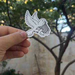 以下が含まれることがあります： 白いダイカットステッカーで、飛んでいる鳩の黒い線画が描かれています。鳥は翼を広げた姿で、くちばしにはフリンジのディテールが施されたケフィエ柄のスカーフをくわえており、背景は白くなっています。