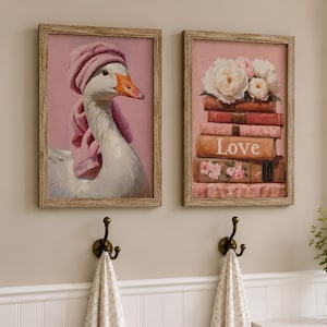 May include: Two rectangular framed wall art prints with light wood frames. The left print shows a white goose wearing a pink towel on its head. The right print features a stack of books with the word Love on the spine, topped with white flowers.