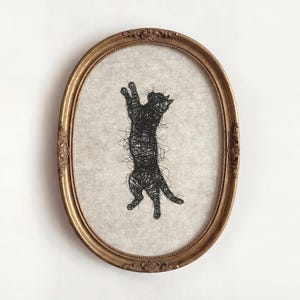 Puede incluir: Una obra de arte enmarcada ovalada dorada que presenta una silueta de gato negro. El gato está representado en una pose juguetona y erguida, creada con un estilo de línea garabateada. El fondo es una tela clara y texturizada.