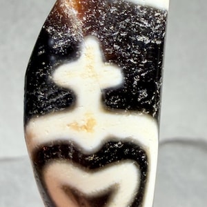 May include: A polished, oblong dzi bead with a distinctive pattern. The bead showcases a central white cross-like design, bordered by black and white bands. The top of the bead has a gradient of brown and white.