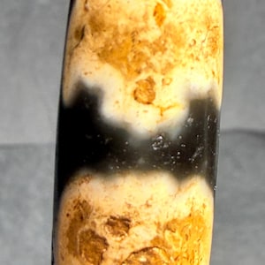 May include: A cylindrical bead with a dark brown top, transitioning to a creamy white and yellow pattern, and a black band. The bead has a smooth, polished surface and a slightly irregular shape, suggesting it is handmade.
