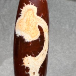 May include: A brown and cream-colored agate bead with a unique pattern. The bead has a cylindrical shape with a rounded top and bottom. The cream-colored design resembles a stylized figure.