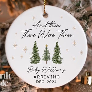 May include: White ceramic Christmas ornament with a green tree design and the text "And then there were three" and "Baby Williams Arriving DEC 2024".