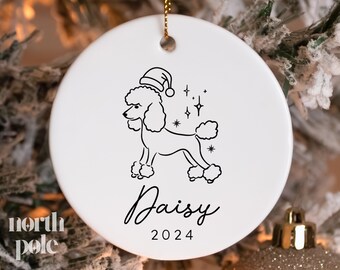 May include: White ceramic Christmas ornament with a black line drawing of a poodle wearing a Santa hat. The ornament has the text "Daisy 2024" on it.