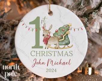 May include: A white round Christmas bauble with a green number one and the word "1st" above it. The bauble also features a green reindeer pulling a small green Christmas tree in a sleigh. The text "CHRISTMAS" is written in green below the number one, and the name "John Michael" is written in red below the text "CHRISTMAS". The year "2024" is written in green below the name.