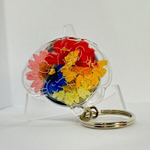 May include: A clear acrylic keychain with a colourful pressed flower design inside. The flowers are red, yellow, blue, and pink.