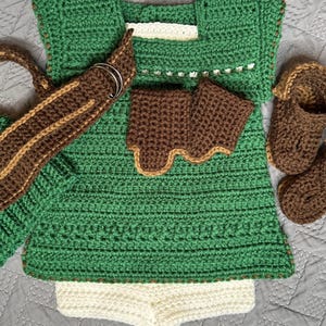 May include: A handmade crochet baby outfit in shades of green, brown, and cream. The set includes a top, shorts, arm cuffs, a belt with silver rings, and booties. The outfit is designed with a textured stitch pattern.