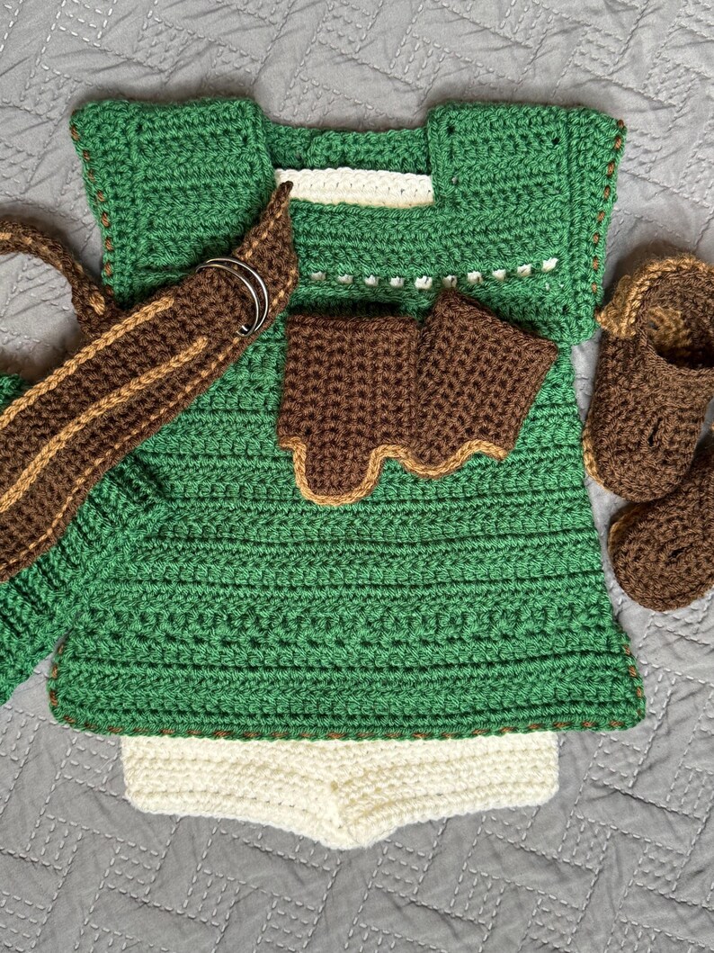 May include: A handmade crochet baby outfit in shades of green, brown, and cream. The set includes a top, shorts, arm cuffs, a belt with silver rings, and booties. The outfit is designed with a textured stitch pattern.