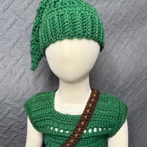 May include: A green crochet costume set, including a hat, tunic, and brown strap. The hat has a pointed tip. The tunic has a square neckline and a brown strap across the chest. The costume is displayed on a mannequin.