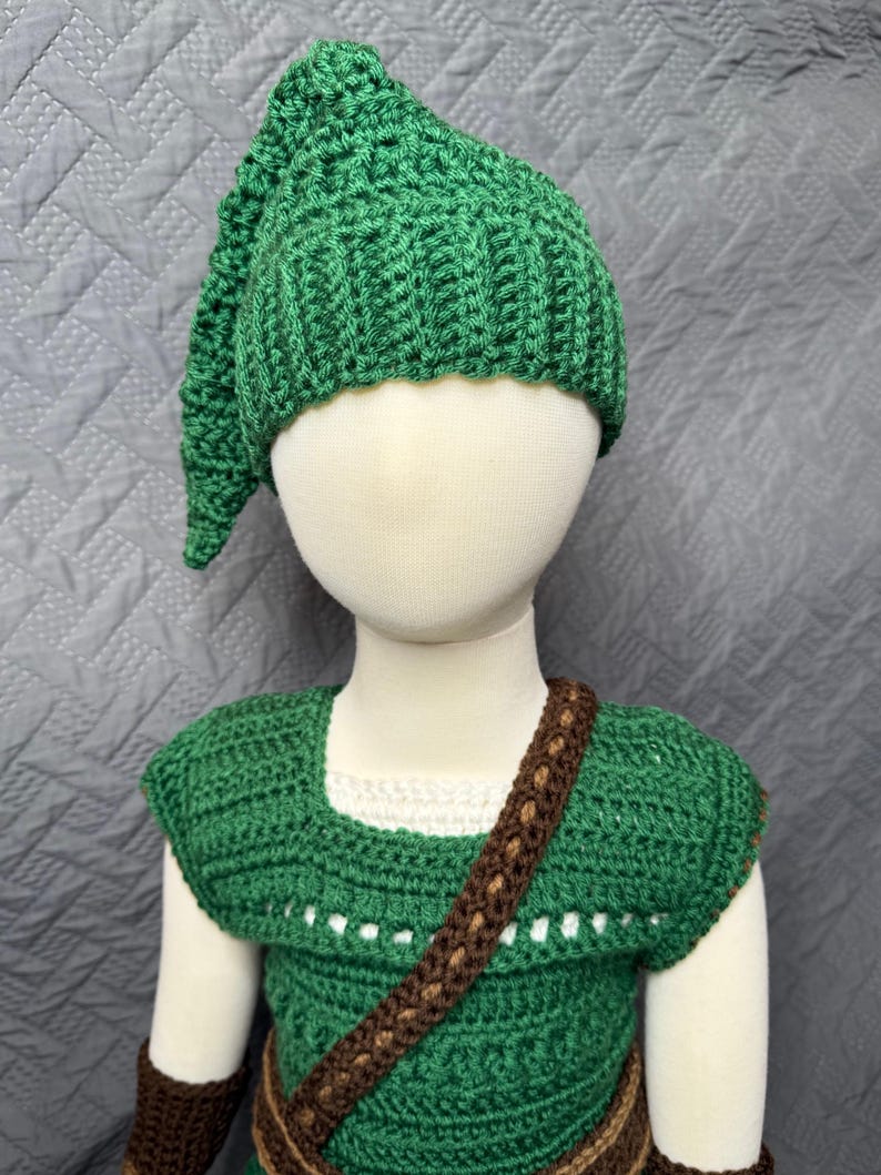May include: A green crochet costume set, including a hat, tunic, and brown strap. The hat has a pointed tip. The tunic has a square neckline and a brown strap across the chest. The costume is displayed on a mannequin.